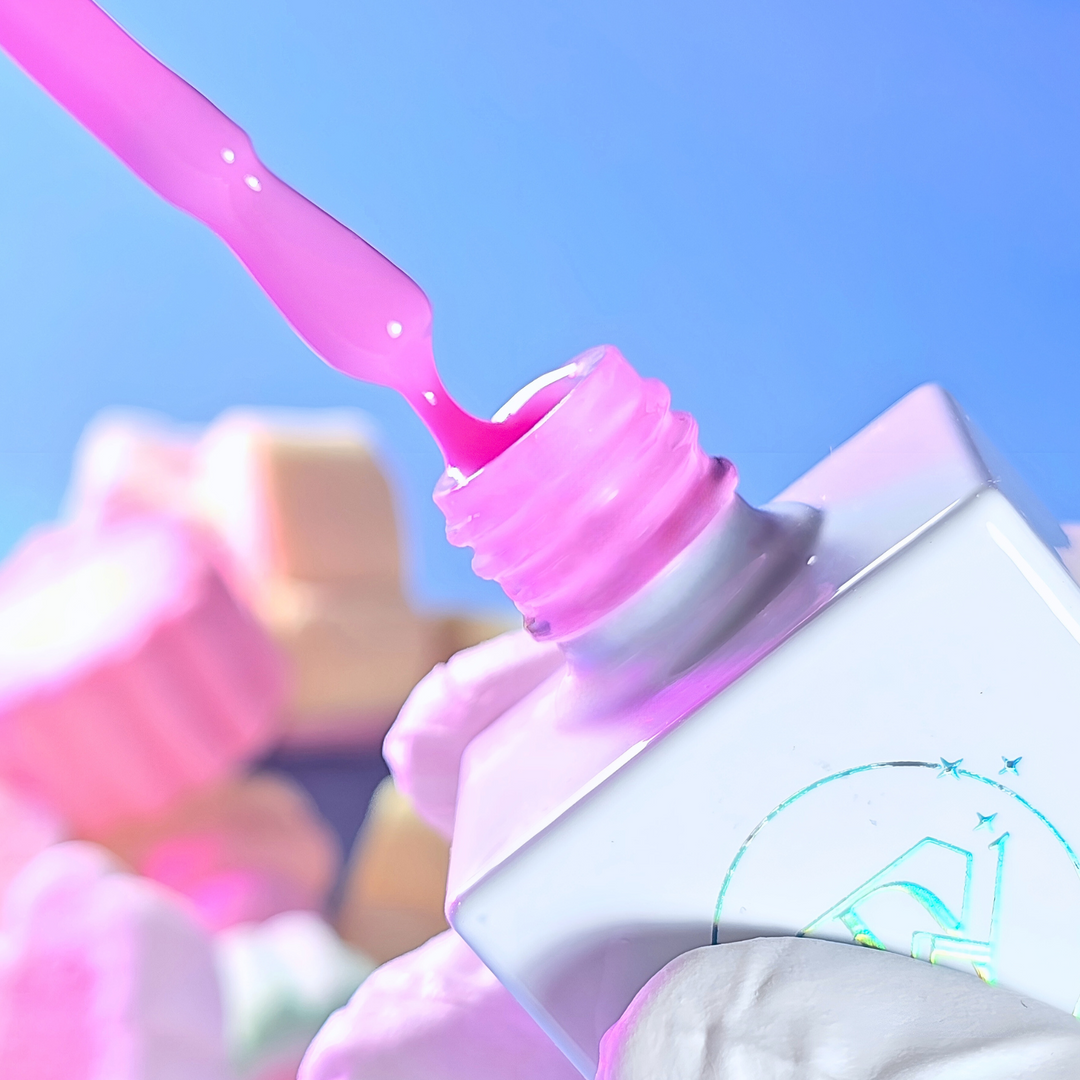 A pink candy-scented base gel being poured from the brush onto the bottle, set against a pastel background with soft lighting.
