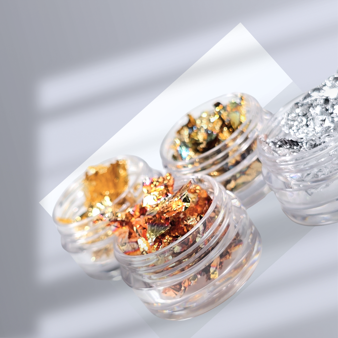 Small clear jars filled with gold and silver foil flakes for nail art, placed on a reflective surface. Perfect for adding metallic accents to nail designs.