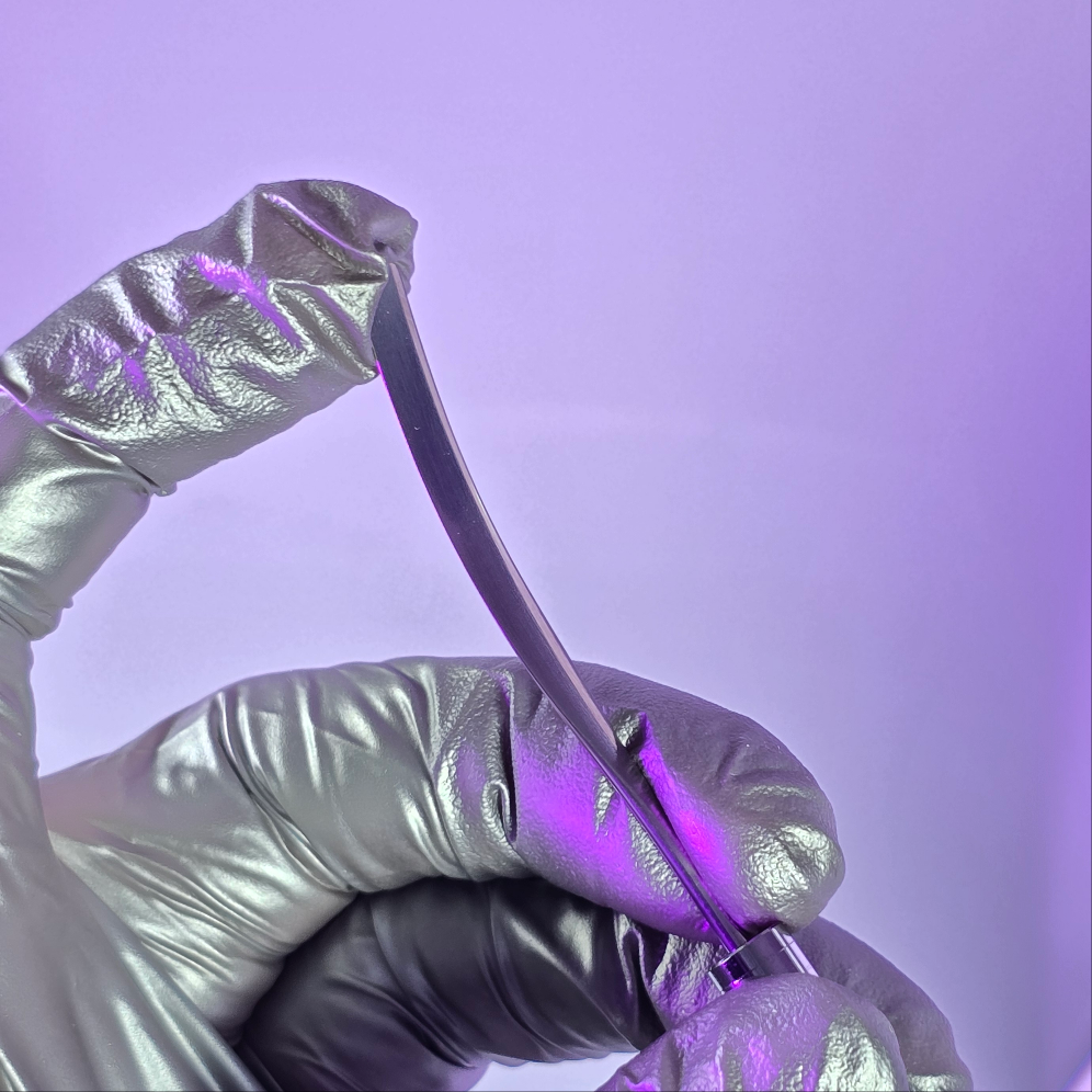 A professional nail artist wearing silver gloves holds a sleek metal spatula tool against a soft purple background, perfect for mixing and applying nail products.