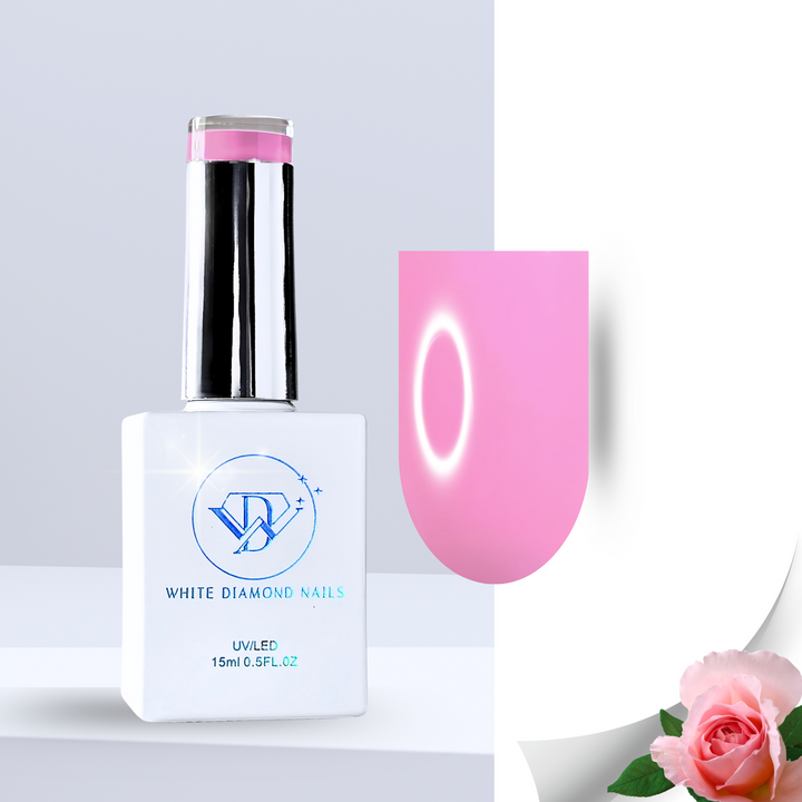 A soft blush pink gel polish swatch on a clear nail tip, reflecting light for a high-gloss finish. The background features delicate pink rose petals, fresh green leaves, and smooth white pebbles submerged in water, enhancing the luxurious, rose-scented theme.