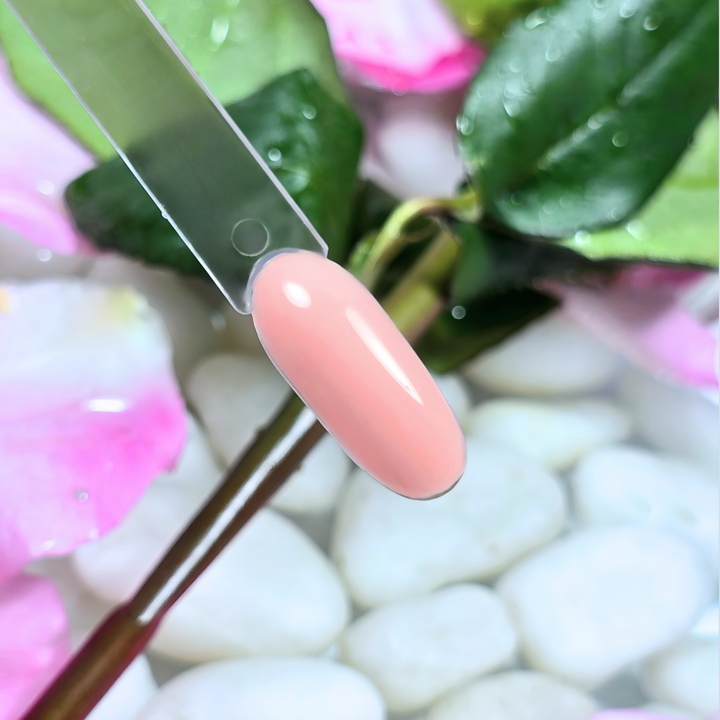 A bold coral pink gel polish swatch with a high-shine, creamy finish, displayed on a clear nail tip. The floral background of fresh green leaves, soft pink petals, and water-drenched white pebbles enhances the vibrancy and energy of this striking shade.