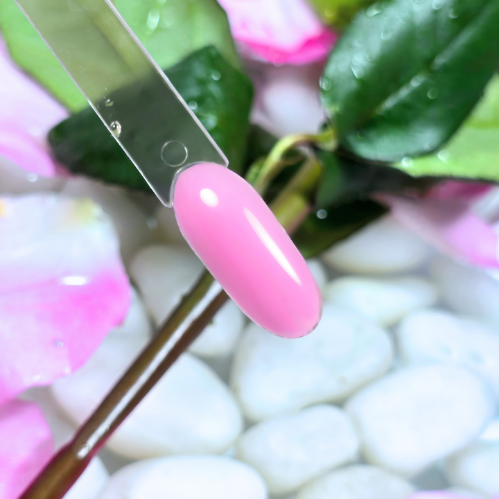 A vibrant bubblegum pink gel polish swatch on a clear nail tip, reflecting light for a sleek, glossy finish. Set against a background of fresh green leaves, pink flower petals, and white pebbles submerged in water, this shade exudes a lively and playful charm.