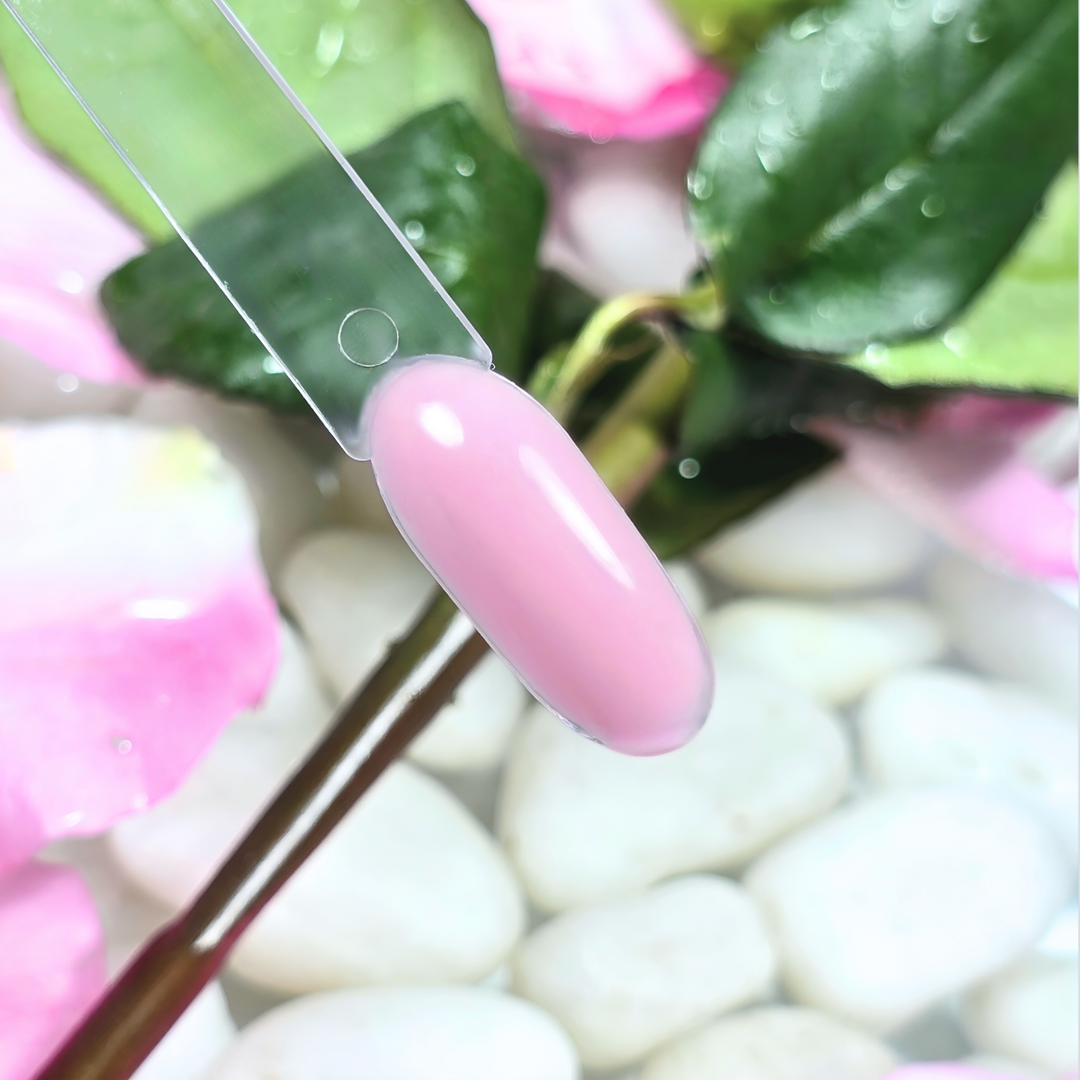 A rich, fresh pink gel polish swatch applied on a clear nail tip, featuring a luminous, high-shine effect. The backdrop of green leaves, pink petals, and white pebbles in water adds to its refreshing, rose-inspired appeal.