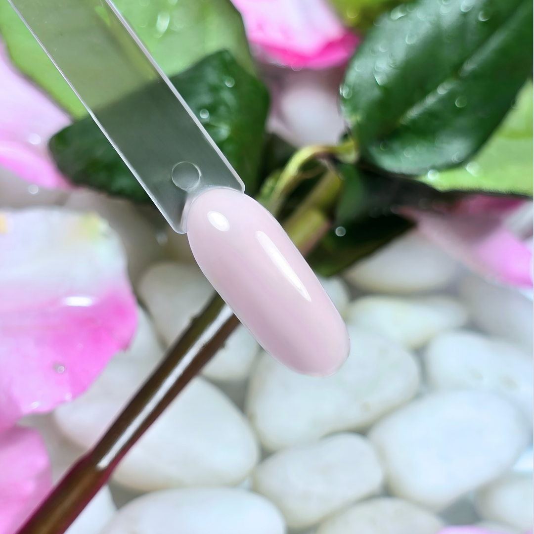 A sheer, milky soft pink gel polish swatch on a clear nail tip with a translucent, dewy finish. Surrounded by lush green leaves, delicate pink rose petals, and smooth white pebbles, this shade embodies natural elegance and subtle beauty.