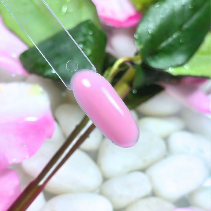 A pastel pink gel polish swatch with a smooth, creamy texture and glossy finish, applied on a clear nail tip. Resting over a floral background of dewy green leaves, delicate pink petals, and white stones, this shade reflects softness and femininity.