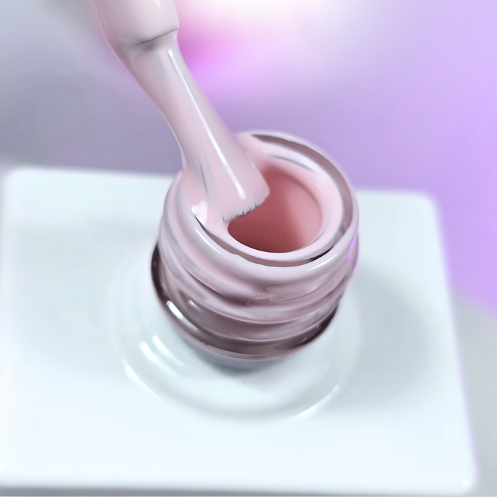 A milky nude pink gel polish being scooped from the bottle with a brush, showing its soft and natural hue. The background features a gentle purple gradient, emphasizing the polish’s neutral and elegant tone.